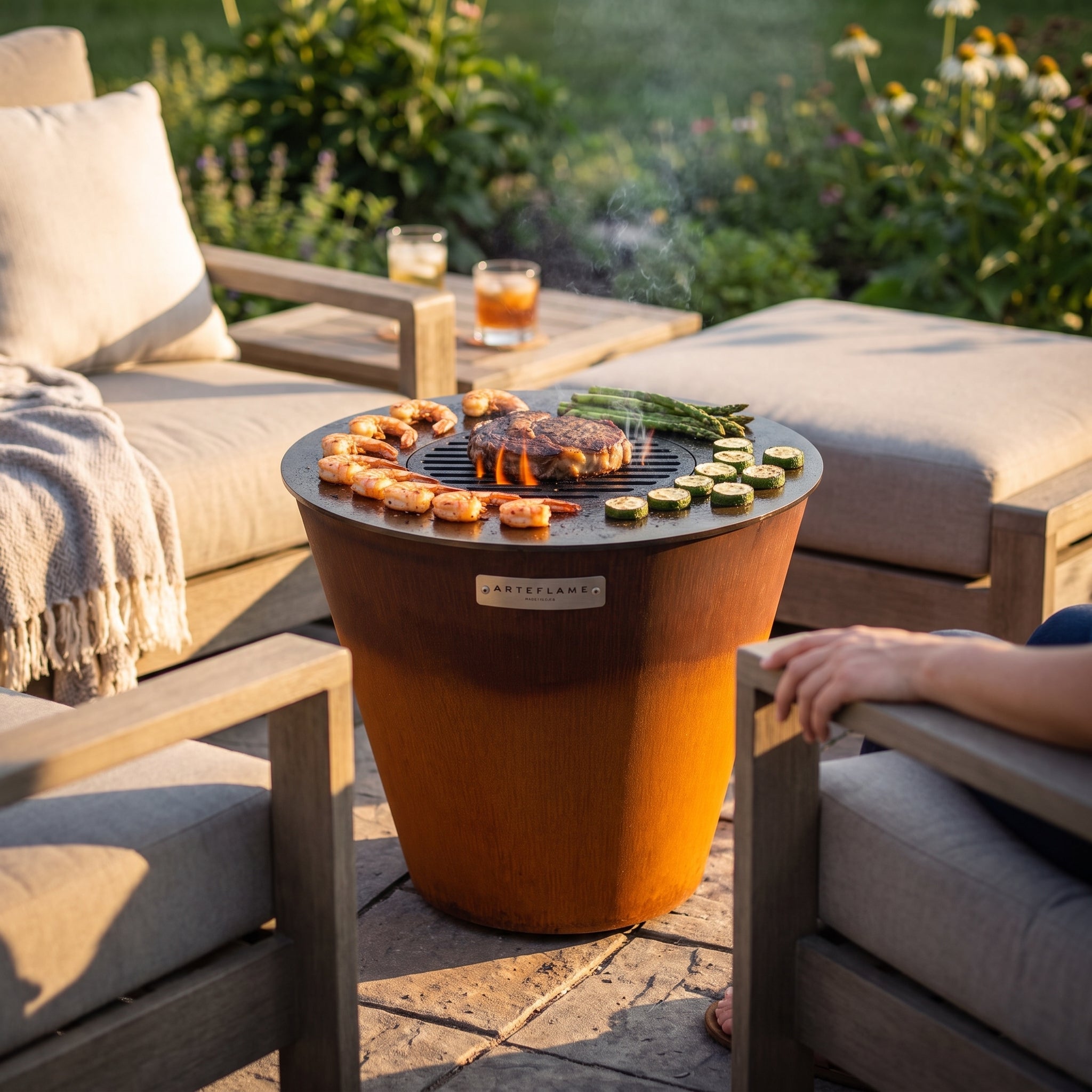 Arteflame ONE20 corten steel grill between two cushioned lounge chairs on a garden terrace, cooking shrimp, steak, asparagus and zucchini at armrest height